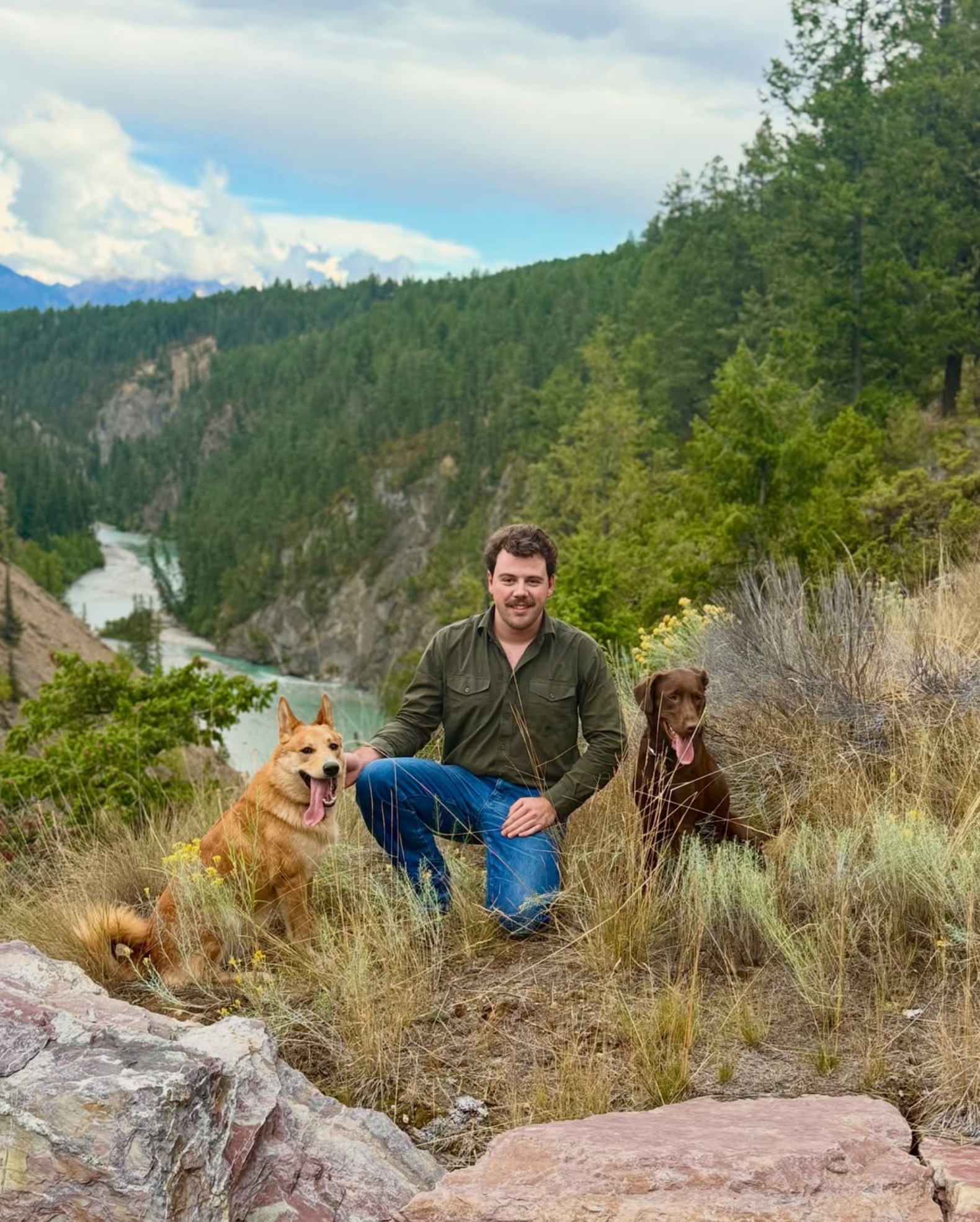 Ryan Neal kneeling beside his two dogs on a hillside overlooking a forested river canyon.